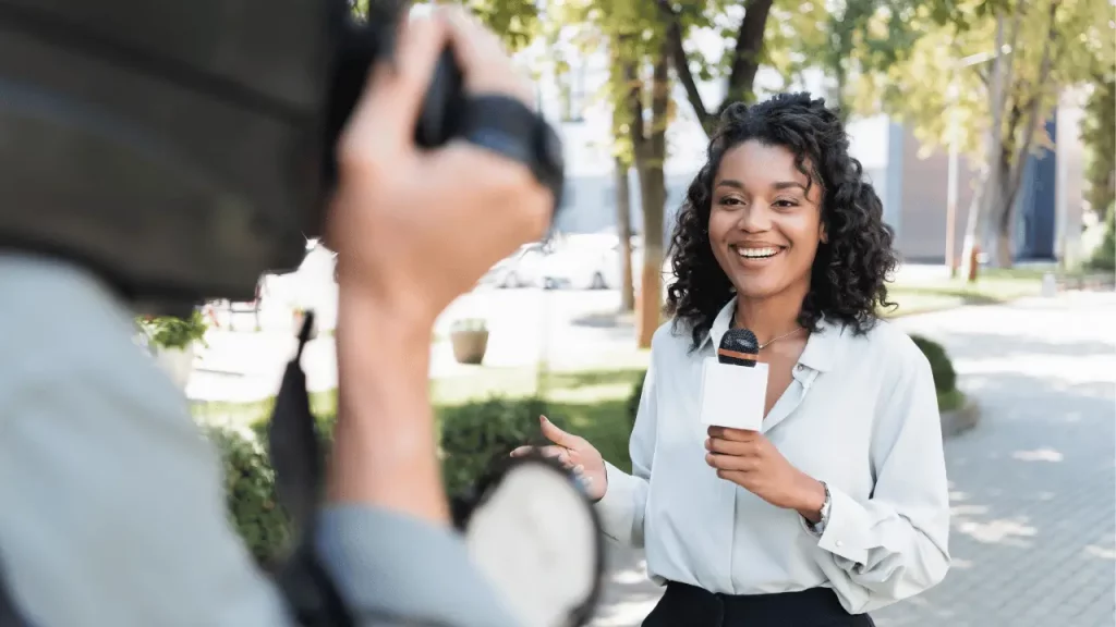 Imagem mostra uma repórter de TV, com o microfone na mão e um câmera filmando ela mostrando a assessoria de imprensa. A mulher é negra e veste camisa social branca.